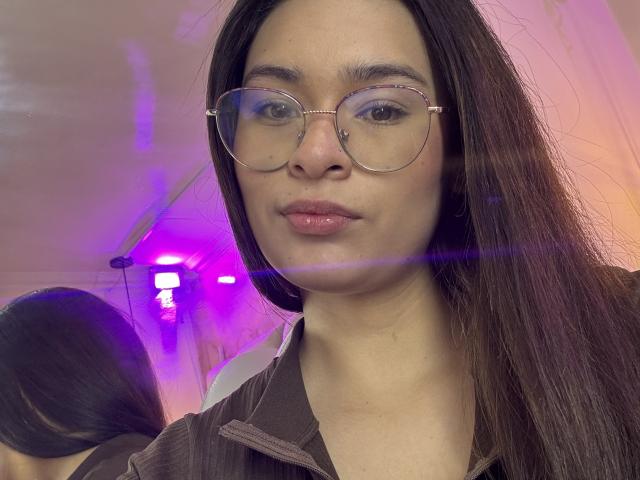 Close-up photo of IsaDoux with long, straight brown hair, wearing round glasses with a small nose stud on the right side. She has a neutral expression and pinkish lips. She's wearing a brown, zip-up jacket. Reflection in a mirror shows her back, highlighting her long hair. Purple and pink light flares are visible across the image. The background is blurred, emphasizing IsaDoux's face and glasses. Her skin tone is light brown.