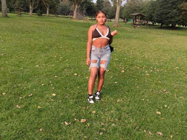 AmberQueeny has brown skin, a slim build, small tits, and wears a white bra, ripped denim shorts, and black sneakers. She stands on green grass, looking at the camera.