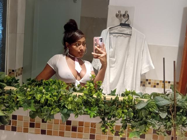 AmberQueeny with dark skin, in a white, deep-cut top showing her large, round tits, taking a selfie in a bathroom with green plant decoration and a white towel.