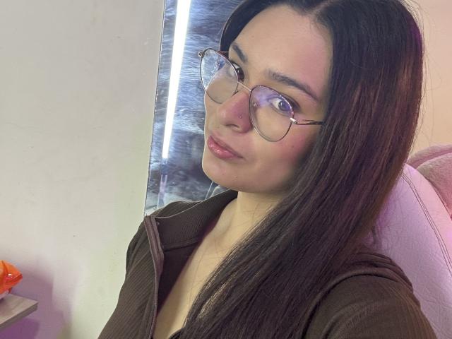 IsaDoux with long brown hair, wearing glasses, and a brown ribbed shirt, pouting slightly, with a soft pink chair in the background. Her face is slightly turned to the camera.