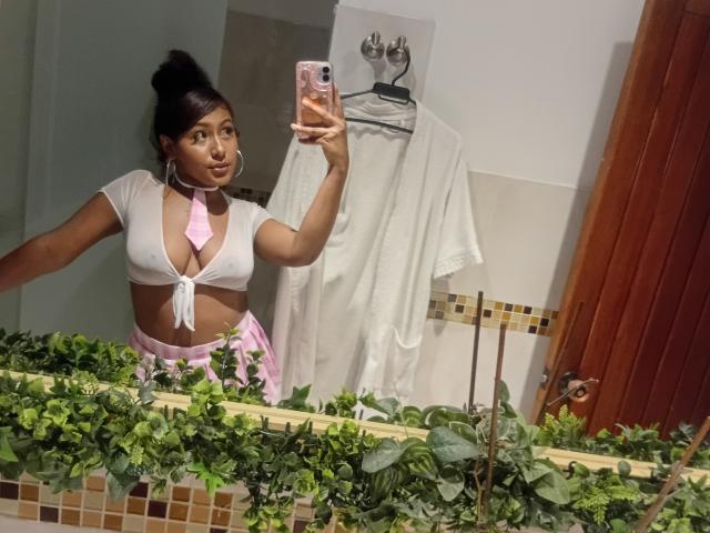 AmberQueeny with brown skin, black hair in a bun, wearing a white tie-top revealing her tits, pink skirt, holding a phone, taking a mirror selfie in a bathroom.