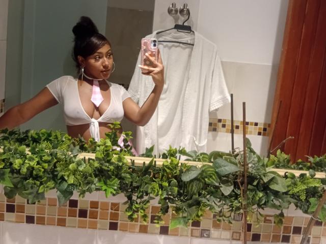 AmberQueeny with dark skin and black hair in a bun, wearing a white, tied-up, short-sleeve top, showing her tits, taking a selfie in a bathroom with green plants.