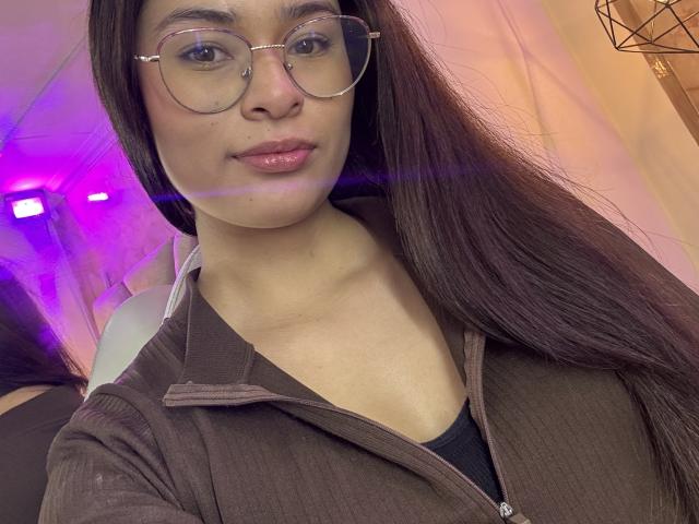 A close-up photo of IsaDoux with long, straight brown hair, wearing round glasses and a brown ribbed zip-up shirt. Her light brown skin is smooth, and she has a slight, confident smile with glossy pink lips. Her black shirt's neckline is visible under the zip-up shirt. The background has purple and pink lights, with a geometric wire frame on the right. The image has a soft, even light. IsaDoux's face is centered, taking up most of the frame.