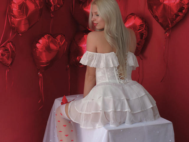 AliceWhitey with long blonde hair, wearing a white off-shoulder ruffle dress, white stockings with red hearts, sitting on a white table, red heart balloons in the background.