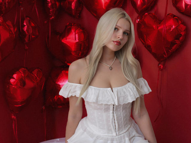 AliceWhitey has long, straight blonde hair and fair skin. She wears a white off-the-shoulder, ruffled dress with a corset-style bodice. Her dress has small, decorative patterns and a slight sheen. She's wearing a silver necklace with a small pendant. Red, shiny heart-shaped balloons are in the background. The composition is centered on AliceWhitey, with a strong contrast between her white dress and the red balloons. The image has a romantic, Valentine's Day vibe.