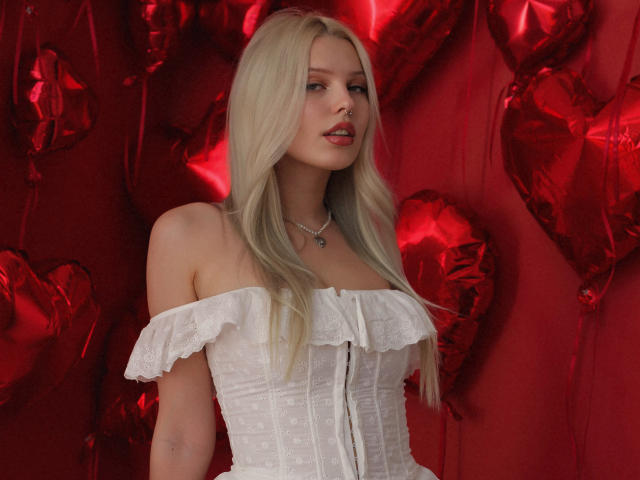 AliceWhitey with long blonde hair, wearing an off-shoulder white lace corset, red lips, and a silver necklace, standing against red heart-shaped balloons. She has small tits.