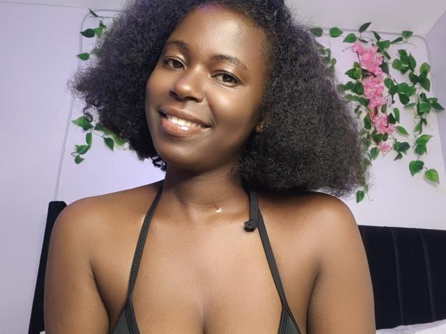 BlackMelanie, avec ses cheveux noirs et bouclés, souriante, portant un haut noir à encolure dégagée, montrant sa petite poitrine, sa peau brune brillante et un joli collier en or..