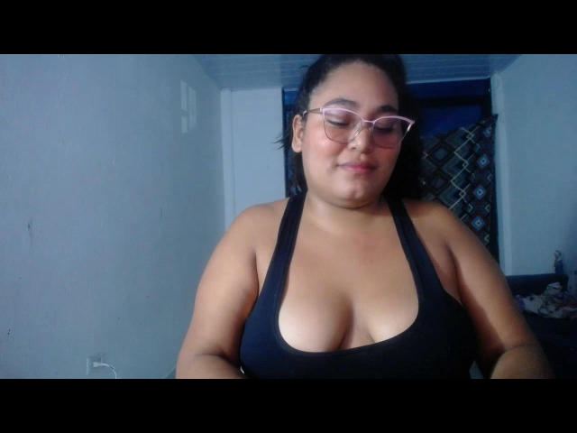 SexyLovve69 with tan skin, black hair, glasses, and a black tank top showing her big boobs, smiling softly in a dimly lit room.