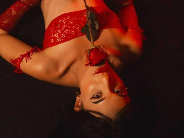 PamelaKander with red lace bra, arms raised, holding rose between her tits, red arm sleeves, black background, red light, dark hair, black eyeliner, seductive, sexy.