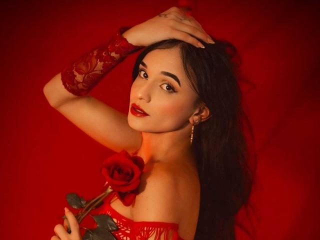 PamelaKander with red lipstick, holding a rose, wearing a red lace glove, and a red off-shoulder top, against a red background, looking seductively at the camera.