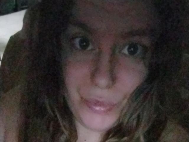 SophiaCoquine has wavy brown hair, large dark eyes, and a slight smile, showing her shiny pink lips. Her face is centered, with soft lighting.