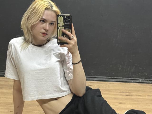 A selfie of SweetLilyy with blonde hair, fair skin, and a slight, relaxed posture. She wears a white, cropped t-shirt with a pink floral design on the shoulder and black pants. Her left arm is raised, holding a phone with a black and gold case, capturing her reflection. She has a small red mark on her lip and a black choker. The wooden floor and dark wall are in the background. Her expression is neutral, slightly pouty. The image is clear, with natural light highlighting her features.