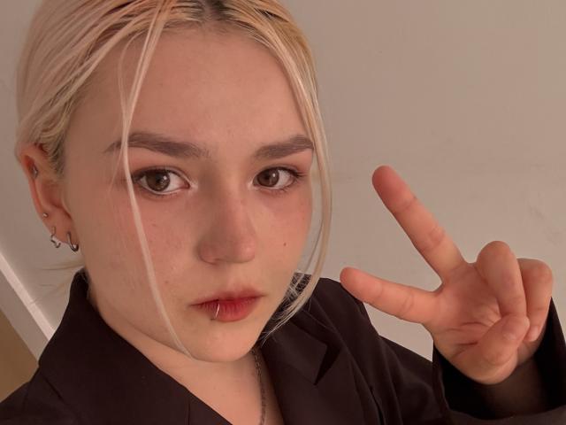 SweetLilyy with blonde hair, brown eyes, and a serious face, making a peace sign with her right hand, wearing a black shirt, and having small piercings in her ear.