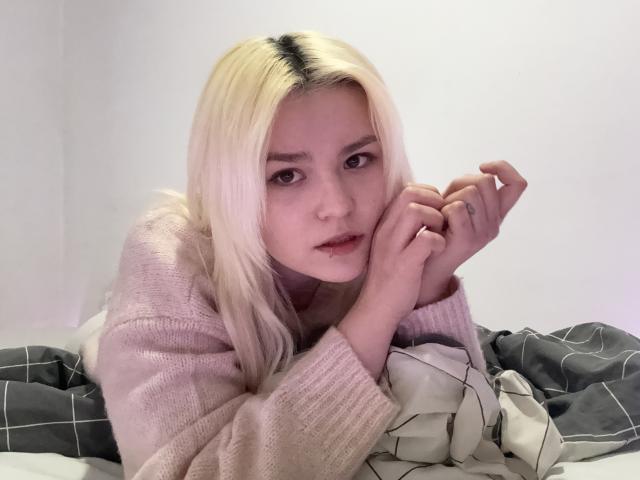 SweetLilyy with pale pink sweater, blonde hair, and white skin, lying on bed, hands near face, looking slightly confused or tired, with gray and white patterned bedcovers.