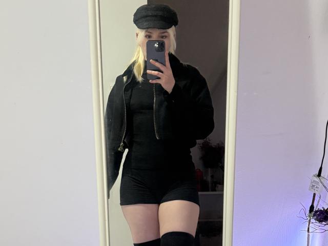 Mirror selfie of SweetLilyy with blonde hair, black cap, black jacket, black dress, black shorts, black thigh-highs, holding phone, showing a slim, curvy body, small tits, ass.