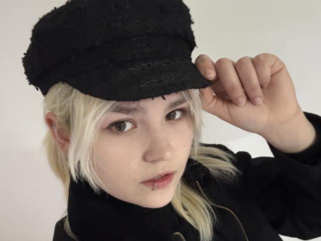 SweetLilyy with pale skin, blonde hair, and a black lace hat, holding it with one hand, wearing a black zip-up jacket, and a lip piercing.