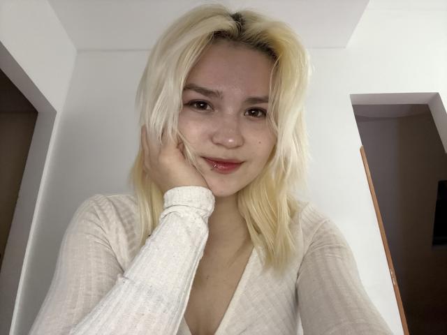 Blond SweetLilyy with fair skin, small nose, and red lips, wearing a white ribbed top, holds her cheek, smiling softly. Her hand touches her face gently.