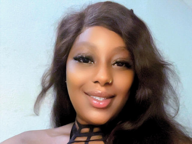 Solene69 has dark brown, wavy hair, a gold nose ring, and is smiling with glossy pink lips. She's wearing a black, strappy top and has long eyelashes.