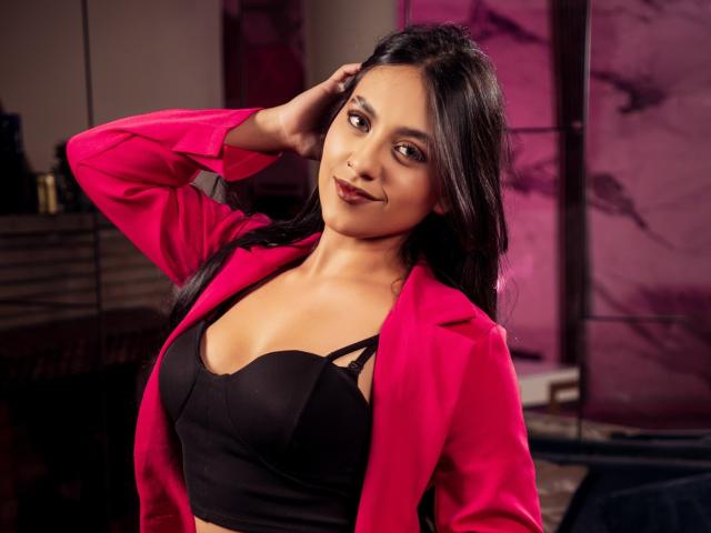 KatyMontclair with long black hair, wearing a pink jacket and black bra, showing small tits, smiling, hand in hair, pink and purple background, looking cute and sexy.