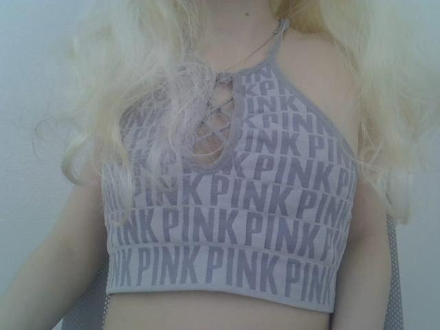 DitaVira wears a gray, patterned crop top with "PINK" in bold letters repeating across the fabric. The top has a lace-up design in the center, exposing a bit of her chest. Her long, wavy blonde hair falls over her shoulders. The image focuses on her upper torso, showing her light skin. The crop top contrasts with her pale complexion. The texture of the fabric is slightly shiny, reflecting light.