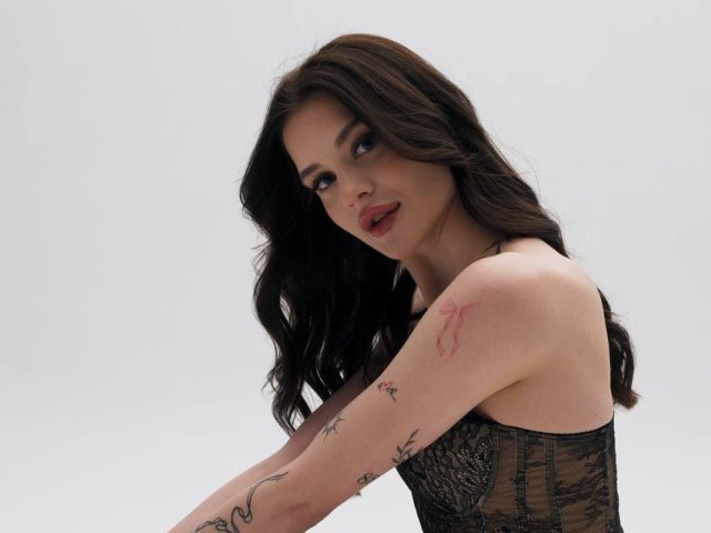 LuisaClover has long, wavy brown hair, dark eyes, and red lipstick. She wears a black lace corset, exposing her tattooed arms. Her tattoos include a red scar-like design on her right upper arm and a small black star on her left forearm. She looks over her shoulder with a slight smile. The image is sharp, with a simple, grey background that emphasizes her figure and tattoos. The composition focuses on her upper body and facial expression. Her makeup is bold, contrasting with her natural hair texture.