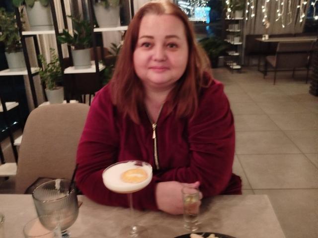 VanessaJuicy with red hair, medium tits, and a slight smile, wearing a red zip-up hoodie, sitting at a table with a cocktail and a glass.