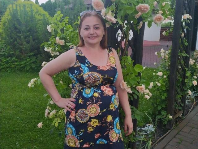 VanessaJuicy with medium tits, wearing a colorful floral dress, standing in a garden with roses, smiling, hand on hip, brown hair, and sunglasses on her head.