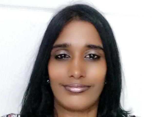 Babrely has dark brown skin, black shoulder-length hair, shiny purple lipstick, subtle eye makeup, and a small smile. Her eyes are dark and she's wearing small earrings.