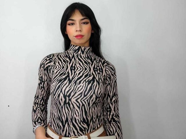 KatherinJohns with zebra-print shirt, long black hair, red lipstick, brown eyes, and white belt, standing against a plain background, looking straight at the camera.