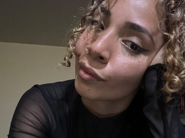 SilvanaXOwen heeft krullend blond haar, een neusring, en draagt een doorschijnend zwart top. Ze kijkt in de camera met een lichtjes fronsende uitdrukking. Haar huid glanst..