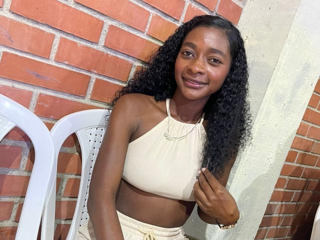 Natix with dark skin, curly black hair, smiling, wearing a beige halter top and matching pants, sitting on a white plastic chair, holding her top with her right hand.