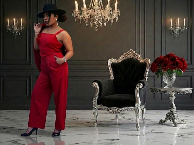SophieSonyas in a red jumpsuit, black hat, and heels, standing with hands in pockets, next to a black velvet chair, silver table with red roses, and crystal chandelier.