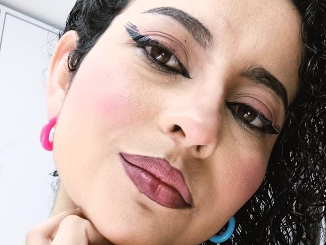 IrinaSweett's face, with pink blush, dark curly hair, thick black eyebrows, pink lipstick, black eyeliner, and pink earrings, looking down with a slight smile.