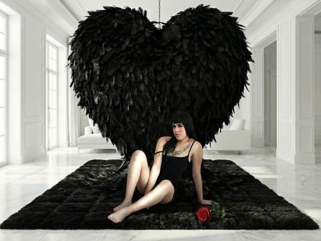 HannaMitsury with black feathered heart, black one-piece, tattooed chest, legs crossed, holding red rose, sitting on black rug, white room, marble floor, black feathers, pale skin.