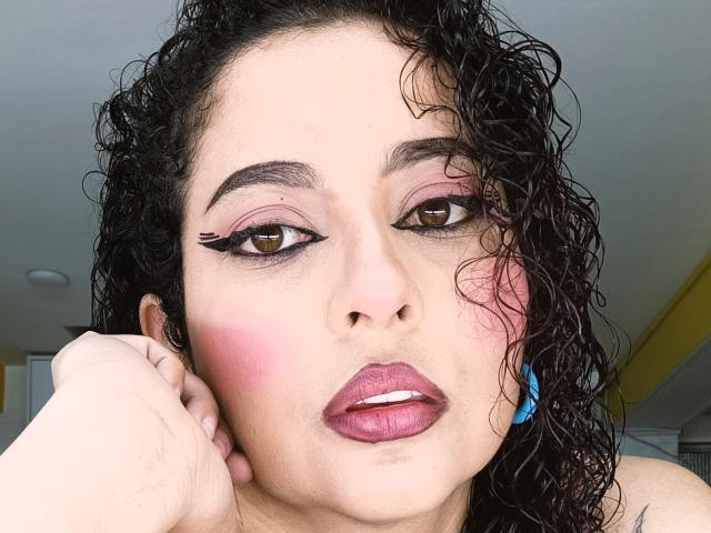 IrinaSweett with curly black hair, brown eyes, pink blush, dark pink lipstick, and blue earrings, holding her cheek, looking at the camera, showing her sexy face.
