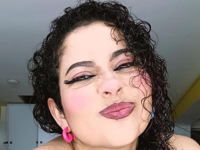 IrinaSweett, met krullend zwart haar, roze lippenstift en roze eyeliner, knipoogt met een fronsende uitdrukking. Ze draagt roze oorbellen en een blauwe oorbeltje. Haar neus is iets omhoog..