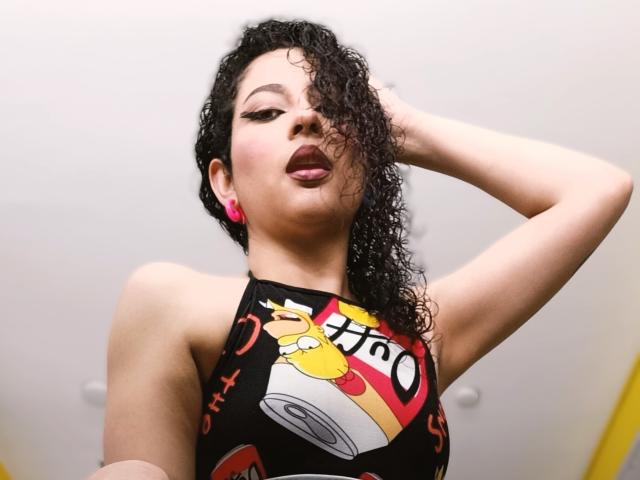 IrinaSweett, avec ses cheveux noirs bouclés, porte un haut noir à encolure américaine avec le logo de Hot Cheetos, des écouteurs roses et du rouge à lèvres. Elle a une main dans ses cheveux et sa bouche est légèrement ouverte..
