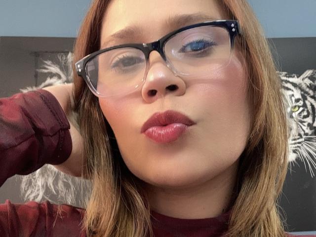 SophiaQueens has glasses, pouty red lips, and a hand in her brown hair. She's wearing a maroon shirt, and her blue eye is slightly closed.
