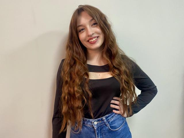 Rioria with long wavy brown hair, black long-sleeve shirt, and blue jeans, smiling, hand on hip, standing against white wall. Boobs are medium-sized, pussy is not visible.