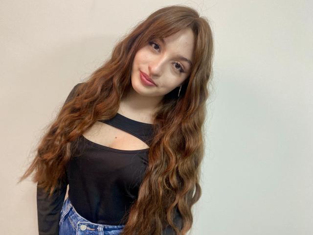 Rioria has long, wavy brown hair, wearing a black top with a cutout, and blue jeans. She's smiling with pink lips, and has fair skin and brown eyes.