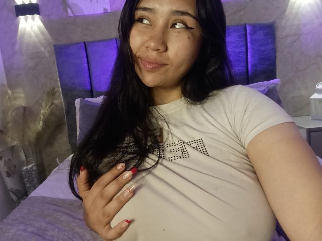 Karollteyes with long black hair, light brown skin, and red nails, smiling at camera, wearing white shirt, hand on her tits, purple-lit bedroom, soft lighting, relaxed expression.