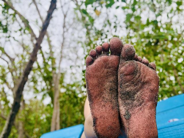 SaraleePoison's dirty, sand-covered soles with her toes pointed upward, showing dirt on her feet, against a blue surface and green, leafy background.