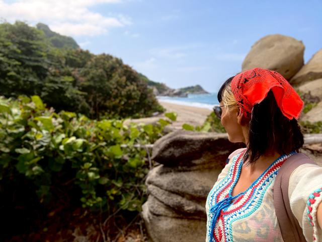 SaraleePoison with red bandana, black hair, wearing colorful top and bead necklace, standing by rocks, looking at beach, has glasses, brown skin, small tits, sunny day.