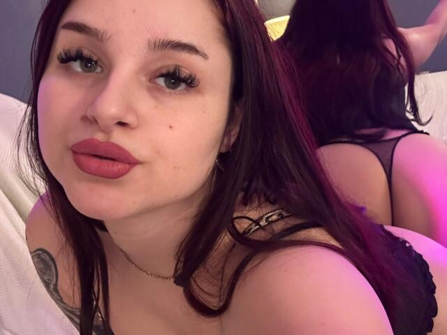 LucieRed with pink lipstick, long dark hair, and a tattoo on her shoulder, wearing black lingerie, looking at the camera while lying on a bed with her ass up.