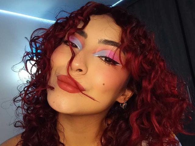 SilvanaGoddess with curly red hair, pink and white eye makeup, and red lips, pouting with a small beauty mark on her cheek, wearing a silver hoop earring.