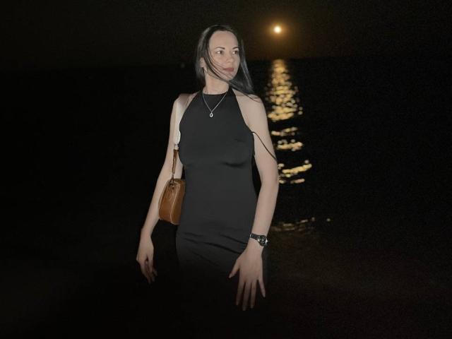 Dalaila with long dark hair, black sleeveless dress, brown leather purse, necklace, and watch, standing by a moonlit water reflection, looking to the side with a slight smile.