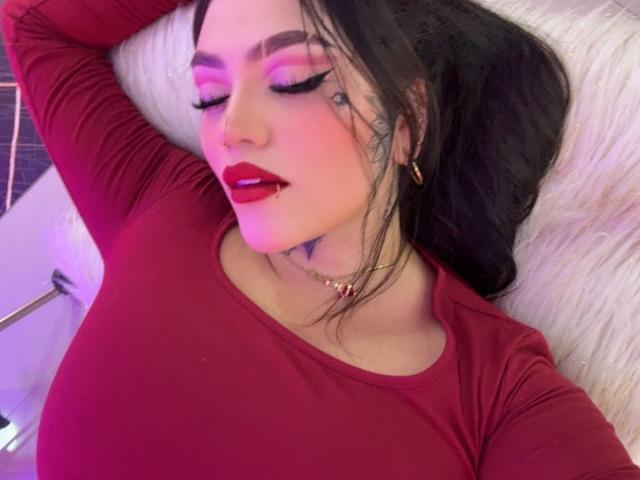 TessaMagic, a dark-haired woman with pink and purple eyeshadow, red lipstick, and a tattoo under her right eye, lies on a white fur pillow. She wears a red long-sleeve top and gold hoop earrings. Her right arm is raised, and her eyes are closed. Her skin is fair, and she has a pink glow on her cheeks. The image has a soft, pinkish-purple light.
