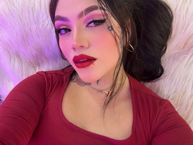 TessaMagic lies on a fluffy white surface. She has long, dark hair, a nose ring, and wears bold red lipstick. Her makeup is dramatic with purple eyeshadow and thick black eyeliner. She has a small tattoo under her right ear. She wears a red long-sleeve top and a gold chain necklace with a red pendant. Her right eyebrow is slightly raised, and she looks directly at the camera with a slightly open mouth.