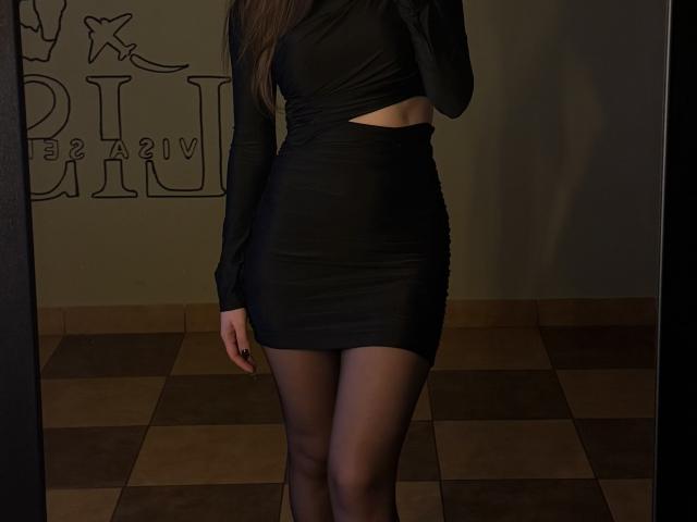 BeccaJoy in a black, long-sleeve, midriff-baring mini dress, black sheer tights, and high heels, showing her ass and thighs. Dark, shadowy room.