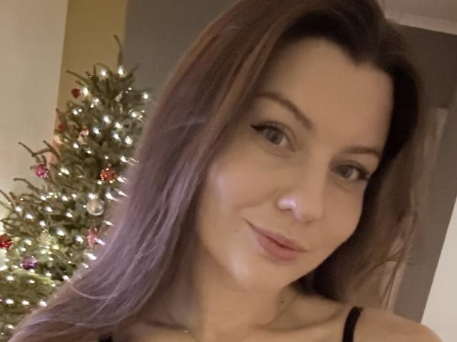 BeccaJoy with long brown hair, subtle makeup, and a slight smile, standing next to a decorated Christmas tree. Her tits are partially visible through a black strap.